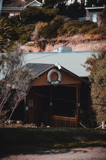 A quaint wooden building nestled among dense greenery features a life preserver with the words 'Canadian Bay Club' displayed prominently at the front. The structure has a small porch with rustic railings, surrounded by trees and shrubs. The background shows a hillside with a mix of vegetation and rooftops of nearby buildings.