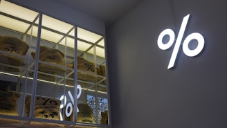 A modern and minimalist interior with a large percentage symbol illuminated on the wall. On the left, shelves display several burlap coffee bags, each labeled with different origins.