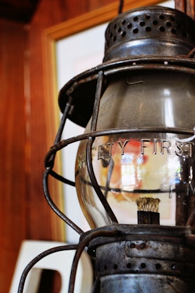 A rustic, metal lantern with a glass enclosure bears the words 'Safety First' etched on its surface. It is set against a wooden background, suggesting an antique or vintage environment.