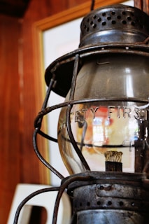 A rustic, metal lantern with a glass enclosure bears the words 'Safety First' etched on its surface. It is set against a wooden background, suggesting an antique or vintage environment.