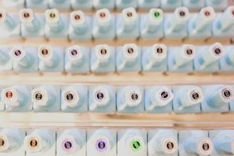 A neatly organized collection of light blue bottles with numbered labels on their tops, arranged in rows. Each bottle has a unique number, with some labels featuring a small colored circle.