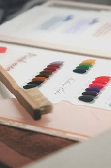 A color palette on paper with a series of vibrant paint swatches arranged in rows, accompanied by a wooden paintbrush resting on the edge. The swatches include an array of colors such as red, purple, green, and orange, neatly organized with labels.