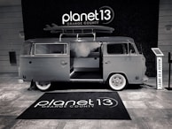 A vintage-style van with a surfboard on its roof is parked indoors. The van has an open sliding door, revealing a modern interior. The floor is covered with a branded rug, and there is a sign encouraging visitors to leave a review.