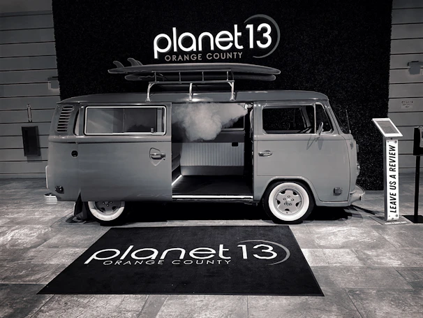 A vintage-style van with a surfboard on its roof is parked indoors. The van has an open sliding door, revealing a modern interior. The floor is covered with a branded rug, and there is a sign encouraging visitors to leave a review.