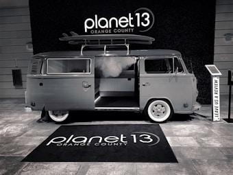 A vintage-style van with a surfboard on its roof is parked indoors. The van has an open sliding door, revealing a modern interior. The floor is covered with a branded rug, and there is a sign encouraging visitors to leave a review.