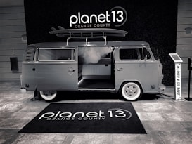 A vintage-style van with a surfboard on its roof is parked indoors. The van has an open sliding door, revealing a modern interior. The floor is covered with a branded rug, and there is a sign encouraging visitors to leave a review.