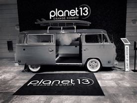 A vintage-style van with a surfboard on its roof is parked indoors. The van has an open sliding door, revealing a modern interior. The floor is covered with a branded rug, and there is a sign encouraging visitors to leave a review.