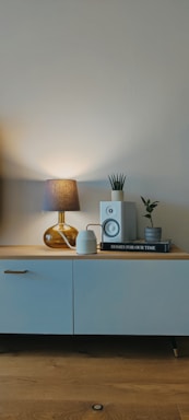 An elegant vintage desk lamp casting a soft glow on a minimalist wooden sideboard with red-orange accent decor.