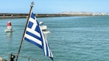 A view of a large body of water with a Greek flag in the foreground. In the background, several small sailboats with red and white sails navigate the calm waters. The scene includes a distant shoreline with rugged, mountainous terrain under a clear blue sky.
