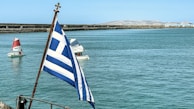 A view of a large body of water with a Greek flag in the foreground. In the background, several small sailboats with red and white sails navigate the calm waters. The scene includes a distant shoreline with rugged, mountainous terrain under a clear blue sky.