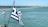 A view of a large body of water with a Greek flag in the foreground. In the background, several small sailboats with red and white sails navigate the calm waters. The scene includes a distant shoreline with rugged, mountainous terrain under a clear blue sky.