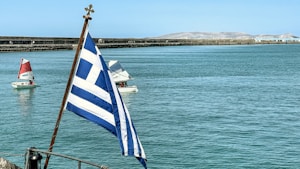 A view of a large body of water with a Greek flag in the foreground. In the background, several small sailboats with red and white sails navigate the calm waters. The scene includes a distant shoreline with rugged, mountainous terrain under a clear blue sky.