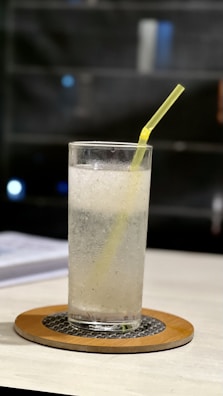 A tall glass filled with a carbonated beverage, possibly soda water, features a yellow straw. It is placed on a table with a coaster underneath. The background is out of focus, suggesting an indoor setting with low lighting.