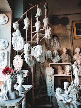 A collection of white plaster busts and sculptures displayed on shelves and a spiral staircase. Among them are various heads in classical style, some featuring hair and facial details, others more simplified. There are flowers placed on some of the sculptures, adding a touch of color. The setting appears to be an art studio or workshop with a rustic, artistic atmosphere.