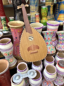 A wooden string instrument is positioned among an assortment of vibrant, painted clay pots and vases. The pots feature various colors and intricate patterns, including floral and geometric designs. The textures and colors create a lively and culturally rich display.