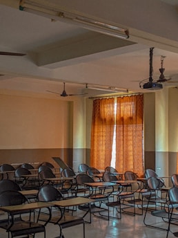 A dimly lit classroom with several rows of empty desks and chairs. The room has beige walls and a brown curtain partially covering the window, letting in some natural light. Ceiling fans and fluorescent lights are visible, along with a mounted projector on the ceiling.