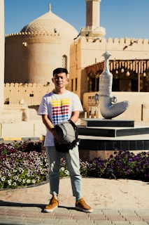 A person stands holding a backpack in front of a historical building with Middle Eastern architectural features, including a domed roof and intricate details. There is a large decorative dagger sculpture on a pedestal nearby. The surroundings include a well-maintained garden with colorful flowers.