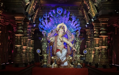 A vibrant and richly decorated idol of Lord Ganesha is placed centrally in an ornate temple setting. The statue is adorned with intricate jewelry, a crown, and a flowing red and white garment. Surrounding the figure are various smaller statues and an elaborate backdrop featuring divine symbols and multiple arms, representing spiritual significance.