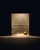 A dimly lit room with plain walls features a vacuum cleaner and a cardboard box placed on the floor. The objects are illuminated by a single overhead light source, creating stark shadows.