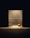 A dimly lit room with plain walls features a vacuum cleaner and a cardboard box placed on the floor. The objects are illuminated by a single overhead light source, creating stark shadows.