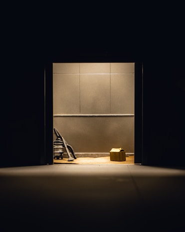 A dimly lit room with plain walls features a vacuum cleaner and a cardboard box placed on the floor. The objects are illuminated by a single overhead light source, creating stark shadows.