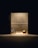 A dimly lit room with plain walls features a vacuum cleaner and a cardboard box placed on the floor. The objects are illuminated by a single overhead light source, creating stark shadows.