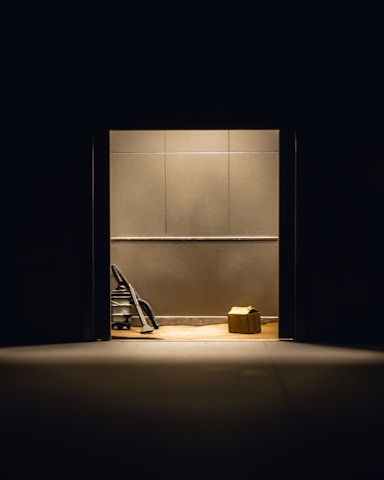 A dimly lit room with plain walls features a vacuum cleaner and a cardboard box placed on the floor. The objects are illuminated by a single overhead light source, creating stark shadows.