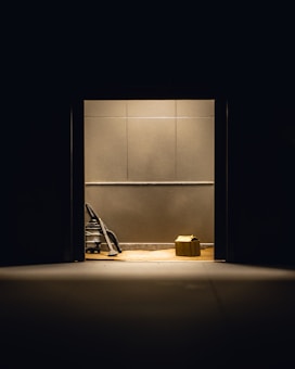 A dimly lit room with plain walls features a vacuum cleaner and a cardboard box placed on the floor. The objects are illuminated by a single overhead light source, creating stark shadows.