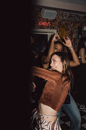 People are dancing and having fun in a dimly lit room with patterned wallpaper and a neon sign that reads 'GIRLS'. The atmosphere appears lively and energetic.