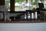 A collection of stainless steel barware including shakers, jiggers, and stirrers displayed on a counter. The background is blurred, indicating an indoor or outdoor bar setting with various objects slightly out of focus.