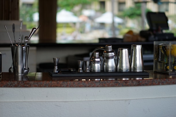 A collection of stainless steel barware including shakers, jiggers, and stirrers displayed on a counter. The background is blurred, indicating an indoor or outdoor bar setting with various objects slightly out of focus.