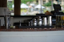 A collection of stainless steel barware including shakers, jiggers, and stirrers displayed on a counter. The background is blurred, indicating an indoor or outdoor bar setting with various objects slightly out of focus.