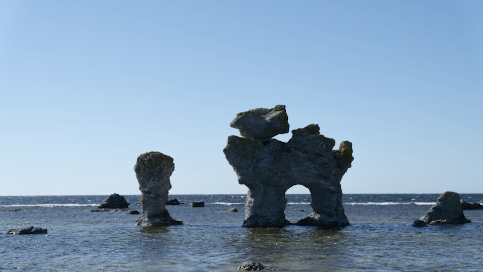 The image depicts a rocky shoreline with three large, gray rock formations standing tall against the backdrop of the ocean. The rocks are arranged in such a way that they appear to be stacked on top of each other, creating an interesting visual effect. The water surrounding these rocks is calm and serene, reflecting the clear blue sky above.