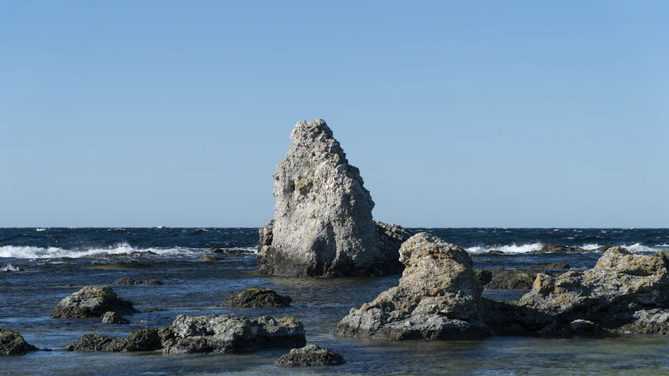 The image depicts a rocky shoreline with a large, gray rock formation standing out against the blue sky and calm ocean waters. The rock formation is situated on the left side of the frame, while the vast expanse of water stretches out to the right. The clear blue sky above adds to the serene atmosphere of the scene.