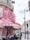 A charming café on a Parisian street, adorned with vibrant pink and red flowers cascading over its awning. The outdoor seating area is occupied by people enjoying the atmosphere. The building features ornate wrought-iron balconies, and a hotel sign is visible above.