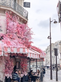 A charming caf&eacute; on a Parisian street, adorned with vibrant pink and red flowers cascading over its awning. The outdoor seating area is occupied by people enjoying the atmosphere. The building features ornate wrought-iron balconies, and a hotel sign is visible above.