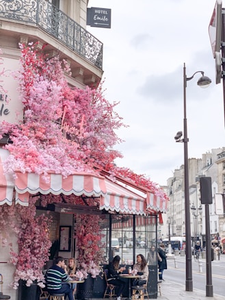 A charming caf&eacute; on a Parisian street, adorned with vibrant pink and red flowers cascading over its awning. The outdoor seating area is occupied by people enjoying the atmosphere. The building features ornate wrought-iron balconies, and a hotel sign is visible above.
