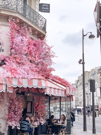 A charming caf&eacute; on a Parisian street, adorned with vibrant pink and red flowers cascading over its awning. The outdoor seating area is occupied by people enjoying the atmosphere. The building features ornate wrought-iron balconies, and a hotel sign is visible above.