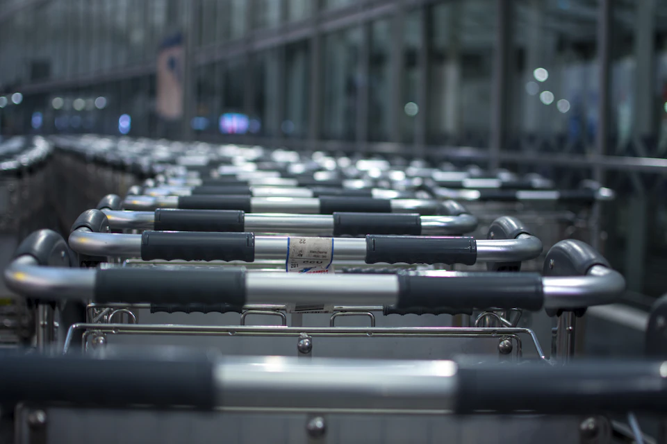 The image shows a row of silver luggage carts lined up against a wall, with each cart equipped with black handles and silver wheels. The carts are arranged in an orderly fashion, creating a sense of symmetry and organization. In the background, there is a large window that allows natural light to illuminate the scene, highlighting the metallic sheen of the carts and their surroundings.