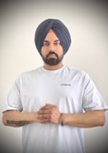A person wearing a dark blue turban and a white T-shirt stands against a plain background. The individual has a beard and a tattoo on their left forearm and wears a metal bracelet on the right wrist. Their hands are clasped together in front.