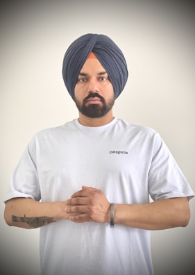 A person wearing a dark blue turban and a white T-shirt stands against a plain background. The individual has a beard and a tattoo on their left forearm and wears a metal bracelet on the right wrist. Their hands are clasped together in front.