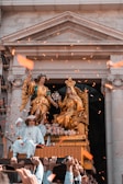 A decorative religious procession with elaborate golden statues depicting a crowned figure seated on a throne and an angelic figure beside them. A child dressed in white, adorned with a crown and angel wings, is sitting in front. The scene is set against the backdrop of a stone building with classical architecture. Orange confetti is being thrown in the air, creating a festive atmosphere.