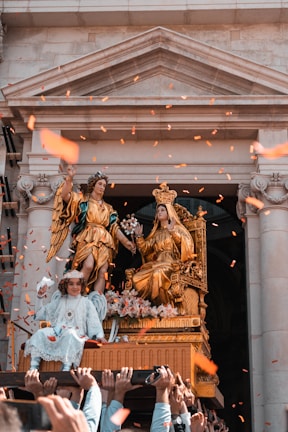 A decorative religious procession with elaborate golden statues depicting a crowned figure seated on a throne and an angelic figure beside them. A child dressed in white, adorned with a crown and angel wings, is sitting in front. The scene is set against the backdrop of a stone building with classical architecture. Orange confetti is being thrown in the air, creating a festive atmosphere.