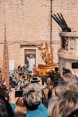 A large crowd of people are gathered around a religious procession. The focus is on a golden religious statue being carried on a ornately decorated platform. Many attendees are capturing the moment with their smartphones. The event takes place in front of a historic stone building, contributing to a vibrant and lively atmosphere.