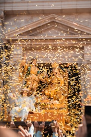 A religious procession features elaborate golden statues adorned with colorful robes amidst a cascade of golden confetti. Several hands are visible, supporting or carrying the platform on which the figures are positioned, capturing a festive and ceremonial atmosphere. The background reveals an ornate architectural structure.