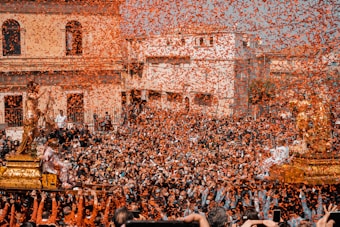 A large crowd of people gathers during a public celebration. The scene is filled with vibrant orange confetti falling from above, adding a dynamic and festive atmosphere. Two ornate statues, likely religious or cultural icons, are being carried on golden platforms. The background consists of historic buildings, suggesting a traditional or cultural event.