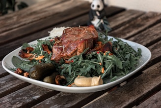 A wooden table holds a plate filled with an appetizing meal. The main focus is a well-cooked piece of steak garnished with rosemary, resting atop a bed of fresh green arugula leaves. Surrounding the steak are slices of mushrooms, small red chili peppers, and a couple of pickles. A light-colored mustard sauce is dolloped on the side. In the background, a small panda figurine is partially visible.