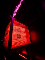 A neon-lit sign features a box with icons and text for drinks, music, coffee, and cake. The background has an overall dark appearance, with prominent red and pink neon lights highlighting the sign.