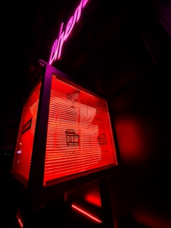 A neon-lit sign features a box with icons and text for drinks, music, coffee, and cake. The background has an overall dark appearance, with prominent red and pink neon lights highlighting the sign.
