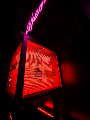 A neon-lit sign features a box with icons and text for drinks, music, coffee, and cake. The background has an overall dark appearance, with prominent red and pink neon lights highlighting the sign.